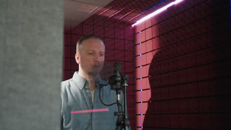 a man wearing a blue long sleeve shirt stands in front of a studio microphone with a straight face, as he stretches his hand in a red soundproofed room