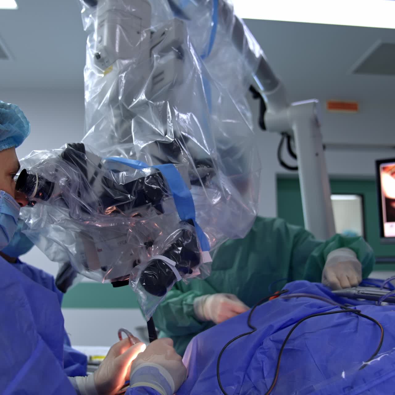 Mid-aged Caucasian surgeon looks intently into microscope performing surgery. Video of operation is shown on the screen at backdrop