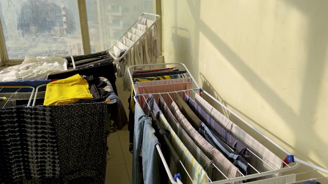Various pieces of clothing hanging on folding drying racks inside a sunlit home. The organized laundry dries naturally near a window during the daytime, representing daily domestic chores and housekee