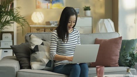 mujer joven con una blusa a rayas y trabajando con una laptop sentada en un sofá en la sala de estar