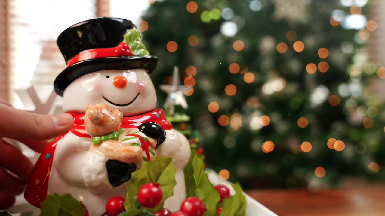 A medium shot of a 5-year-old girl placing ornaments on a Christmas tree. The focus shifts from her to a glass snowman cookie jar in front of the camera in 4k.