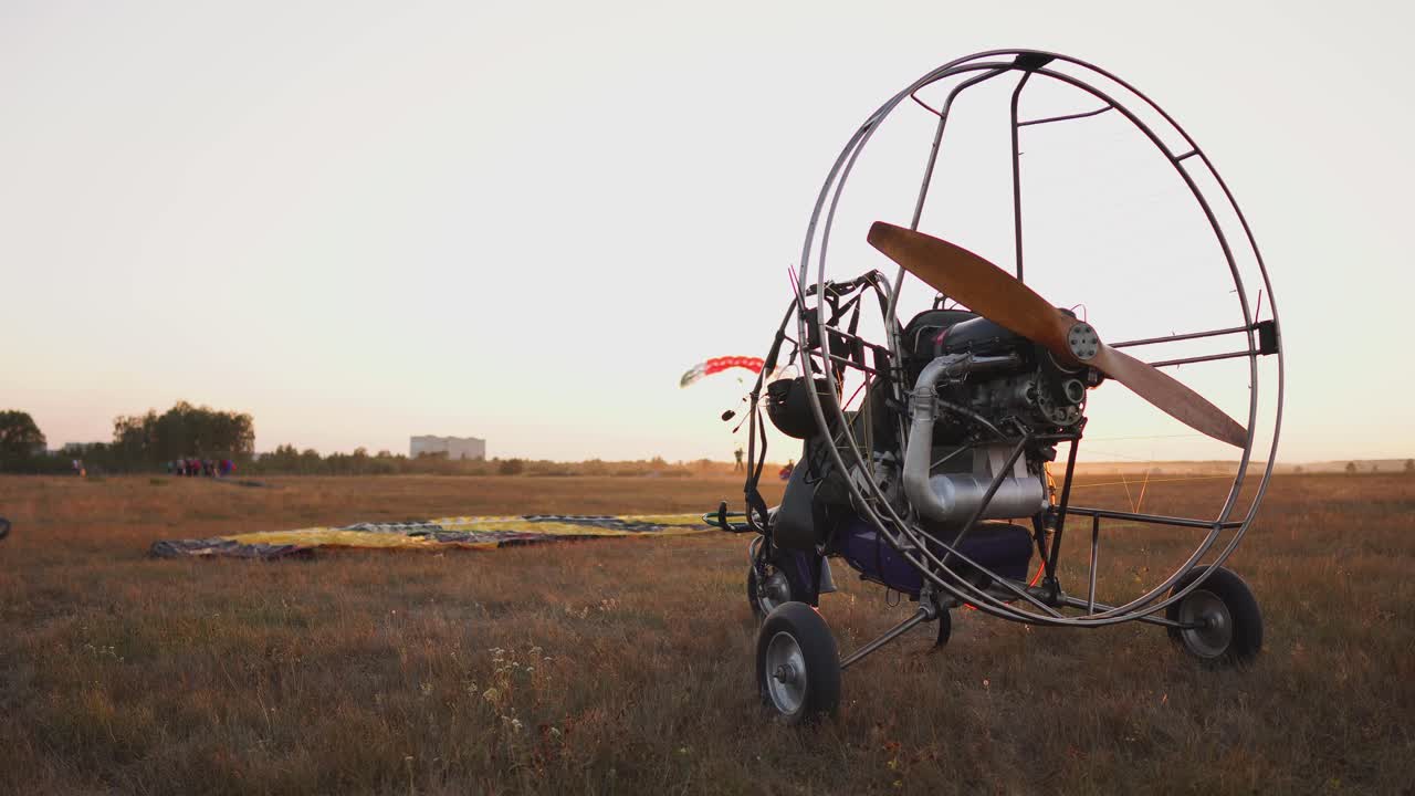 모터 패러글라이더는 해가 지는  ⁇ 빛 아래 공항에 서 있습니다. 배경에는 패러글라이더가 착륙하고 카메라가 서서히 패러글라이더 주위를 움직입니다.