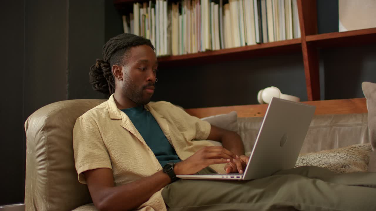 African American man starting typing on laptop on leather couch, pausing to think, bookshelf