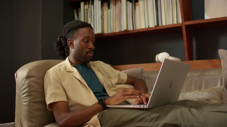 African American man starting typing on laptop on leather couch, pausing to think, bookshelf