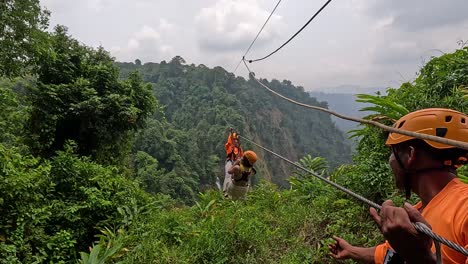 a person wearing safety gear rides a zipline across a dense, green jungle valley under cloudy daylight, with guides assisting at the landing platform. wide shot, natural lighting, steady camera
