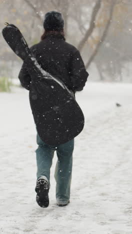 vista trasera de alguien con una gorra negra y una sudadera con capucha con las manos en el bolsillo, llevando una guitarra en su mochila, caminando a través de la nieve, con un banco y árboles en el fondo