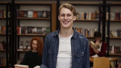 Close-Up-Portrait-Attractive-Young-Man-Laughing-Enjoying-Successful-Lifestyle-Achievement-Cheerful-Male-In-Library-Bookshelf-And-Classmates-On-Background-College-Education