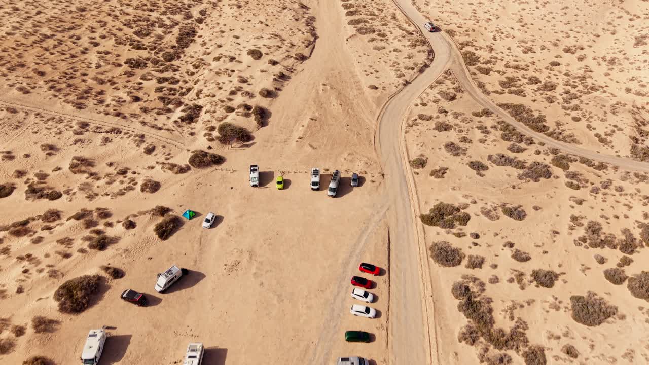 Forward tilt up drone view of a coastal parking area with cars and motorhomes in Fuerteventura, near a volcanic beach. Shot at sunset with golden tones, evoking travel spirit and warm tranquility