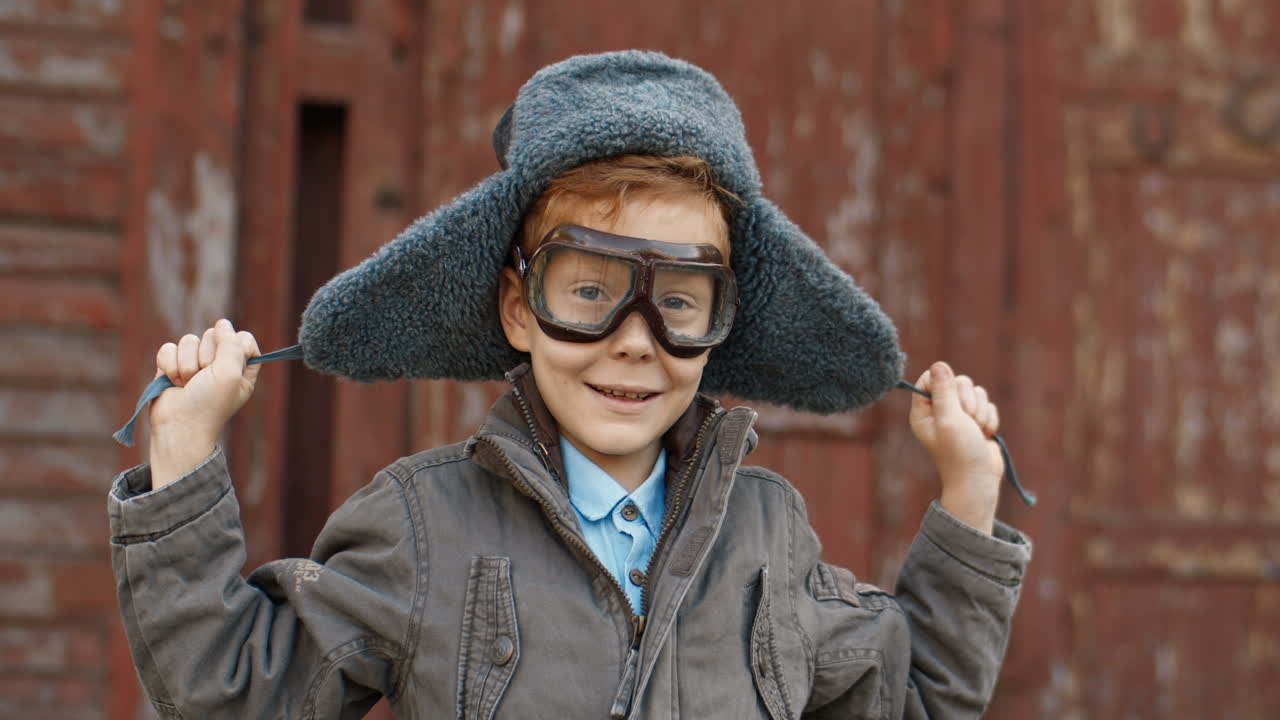 Little Red Haired Boy In Hat And Glasses Playing To Be An Aviator While Looking And Smiling At The Camera