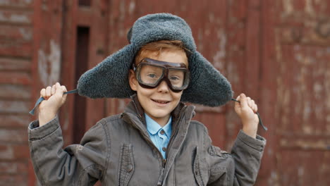 little red haired boy in hat and glasses playing to be an aviator while looking and smiling at the camera