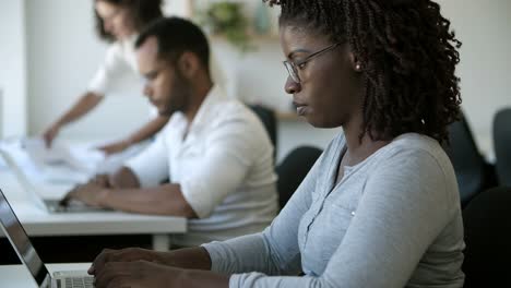 side view of focused female worker in eyeglasses typing on laptop.