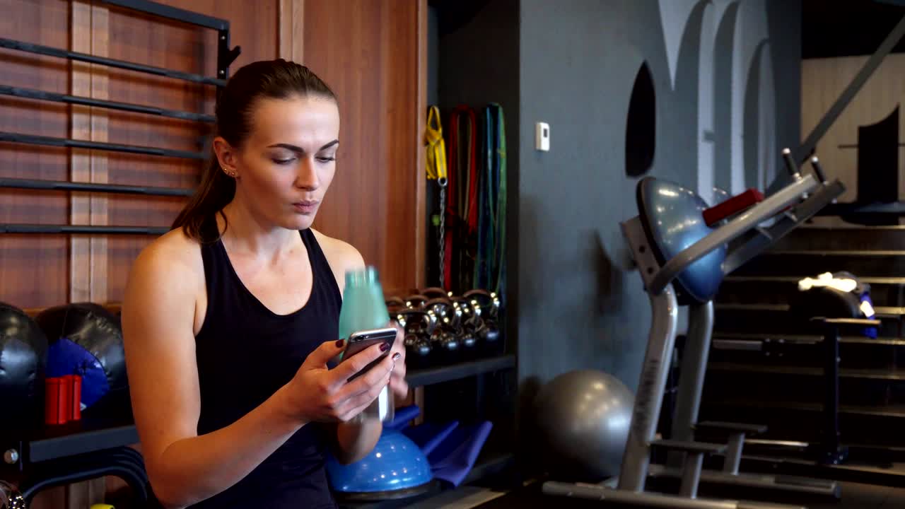 la chica deportiva mira el teléfono y bebe agua durante un descanso entre los ejercicios