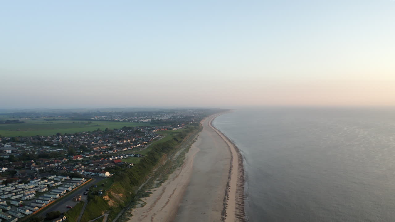 일몰 때 영국 노퍽 해안의 풀이 무성한 평평한 땅이 있는 긴 모래 빈 해변을 따라 날아가는 드론 공중 촬영