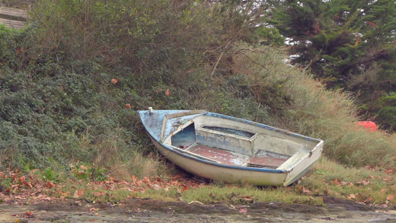A small, old wooden rowboat with heavily worn paint lies abandoned in the grass and dead leaves. The scene evokes time passing and quiet rural decay.