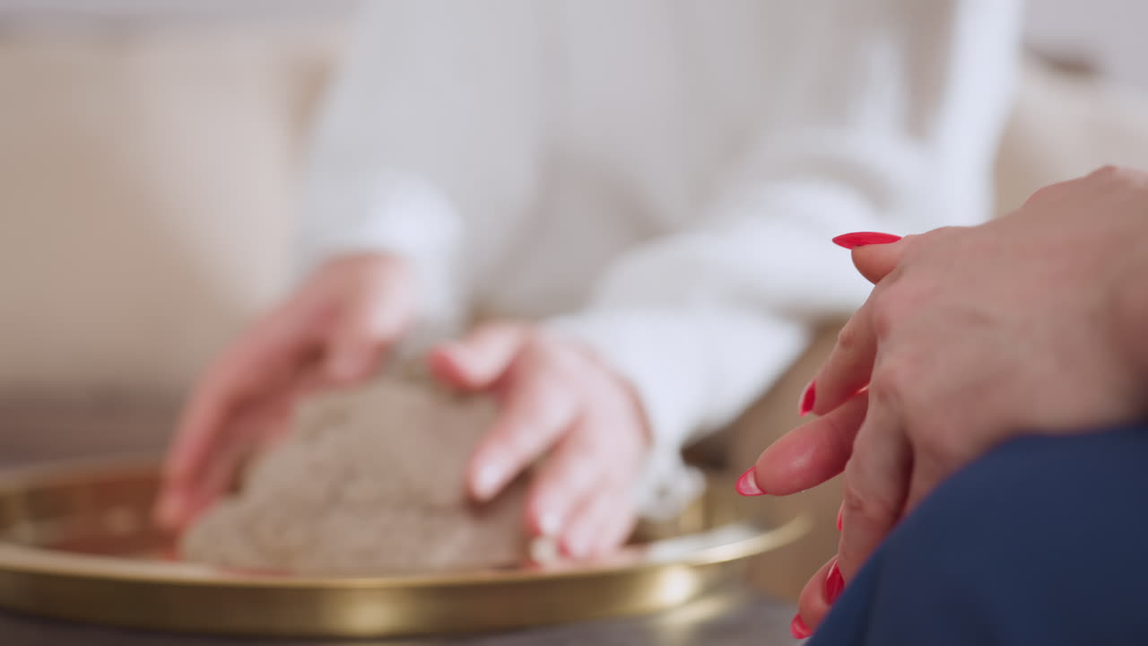 Red manicured hands of specialist in foreground observing participant gently lifting soft wet soil mold during therapy session, capturing moment of tactile interaction in calm indoor setting