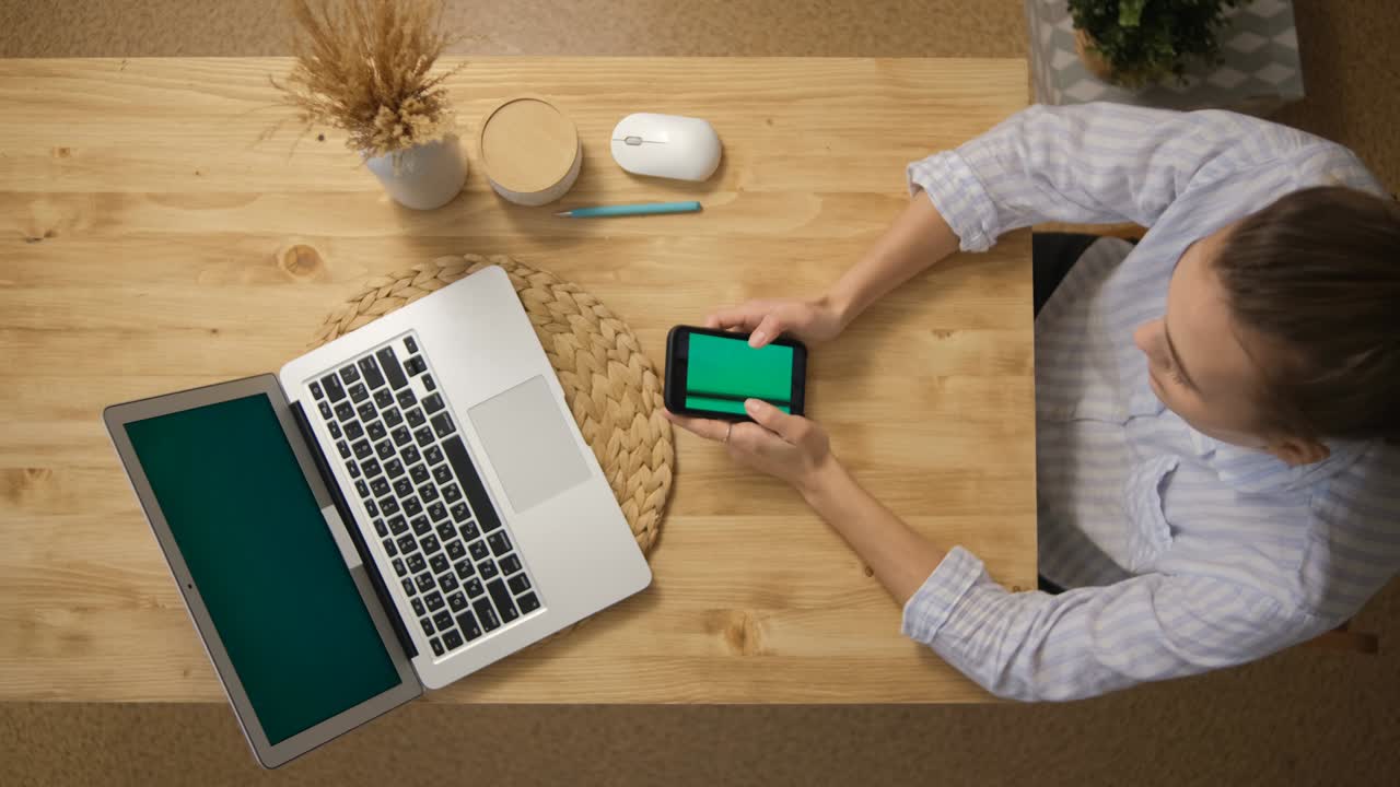 The girl is surfing the Internet. and sits in social networks instead of remote work and conferences. The green screen of the phone monitor. A laptop with a green screen. Top view. Remote work.
