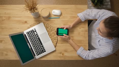 the girl is surfing the internet. and sits in social networks instead of remote work and conferences. the green screen of the phone monitor. a laptop with a green screen. top view. remote work.