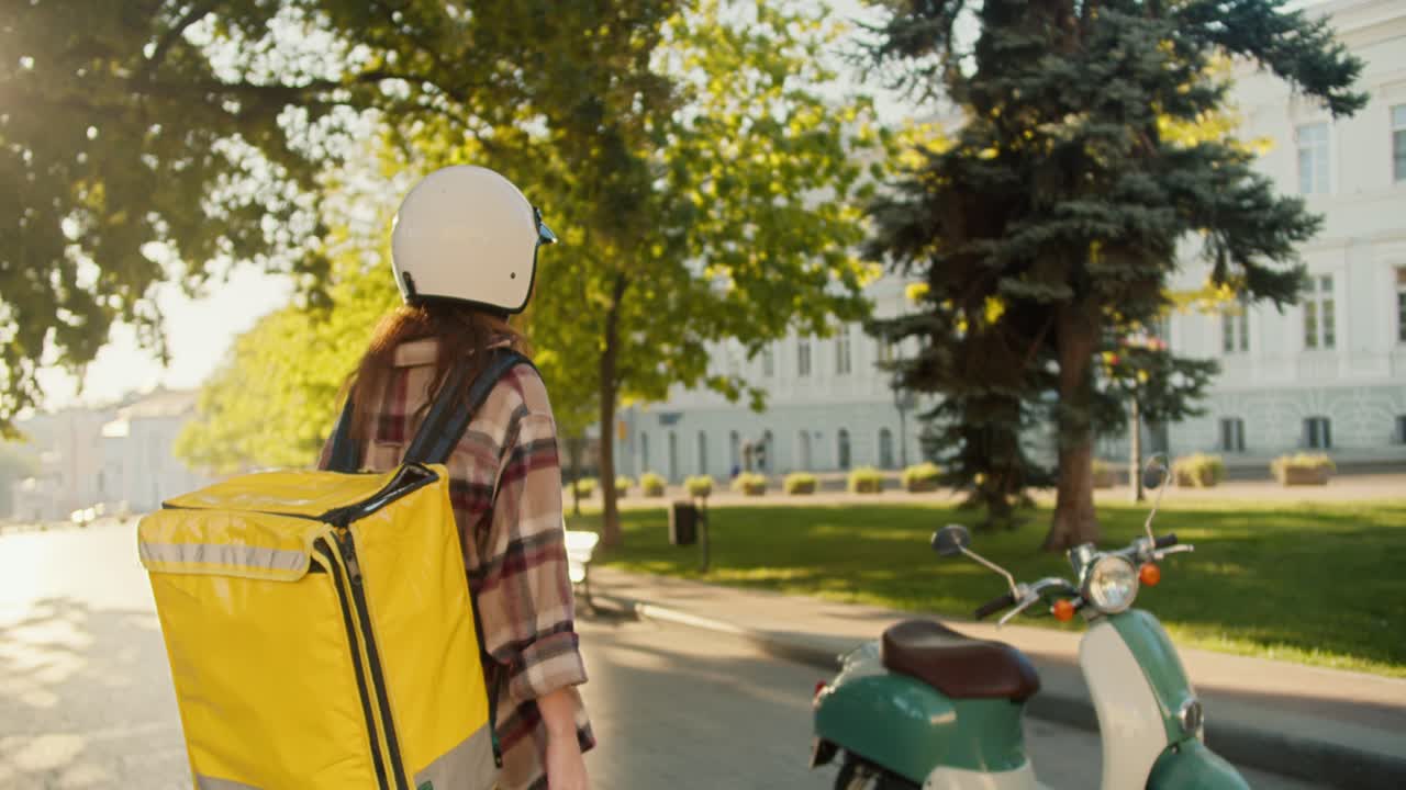 Rear view of a girl courier in a checkered shirt in a white helmet and with a large yellow bag goes to her moped, gets on it and starts moving to bring the order on the street near the park in sunny weather in the summer