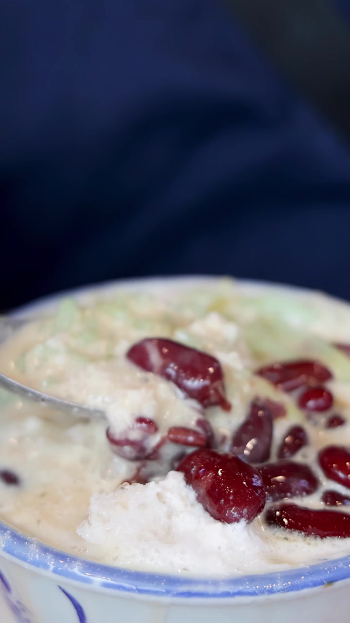 Sendok logam menyendok es cendol dengan kacang merah, es serut, dan santan dalam mangkuk di bawah pencahayaan lembut dan merata dengan perspektif jarak dekat.