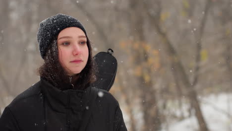 lady in black hoodie carrying a guitar with hands in pockets walking thoughtfully under snowflakes, snowy bench in background adds to serene winter atmosphere