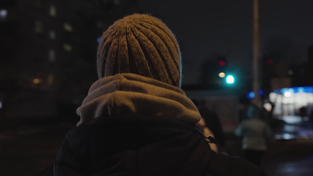 L'inseguimento da dietro segue un uomo con felpa con cappuccio a strati e berretto che cammina nel paesaggio urbano del centro di notte.