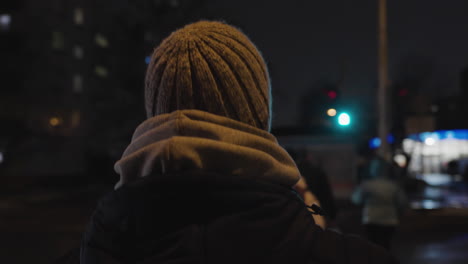 el seguimiento de la vista trasera sigue al hombre con sudadera con capucha en capas y gorra caminando por el centro de la ciudad por la noche