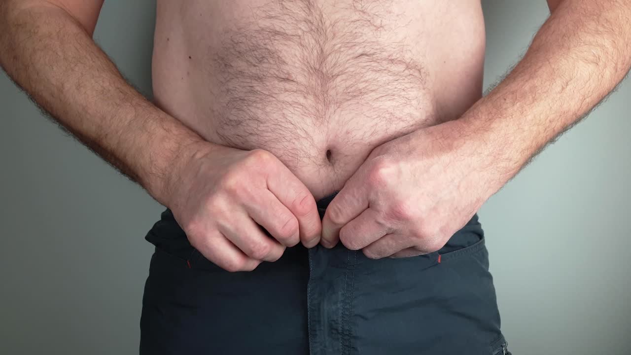 un hombre sin camisa con sobrepeso tirando de su estómago para entrar en los pantalones