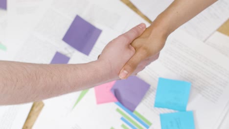 Overhead-Close-Up-Of-Business-Colleagues-Shaking-Hands-Over-Paperwork-On-Table