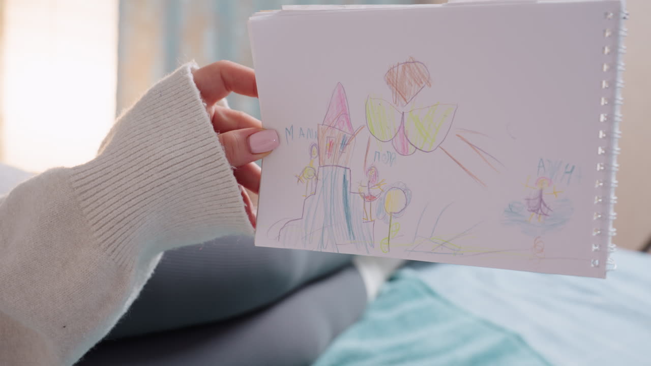 Close up of hands holding sketchbook with colorful child drawing of house, people, tree, sun, pointing finger showing detail, cozy home setting with natural light and soft background atmosphere