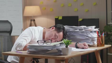 지친 아시아인 남자가 사무실에서 문서와 함께 열심히 일해서 잠을 자고 있습니다.
