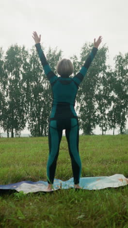 vista trasera de una mujer de pie con la cabeza ligeramente levantada, levantando las manos lentamente extendidas, luego bajándolas cerca de su cara mientras practica yoga en un vasto campo abierto bajo un cielo nublado