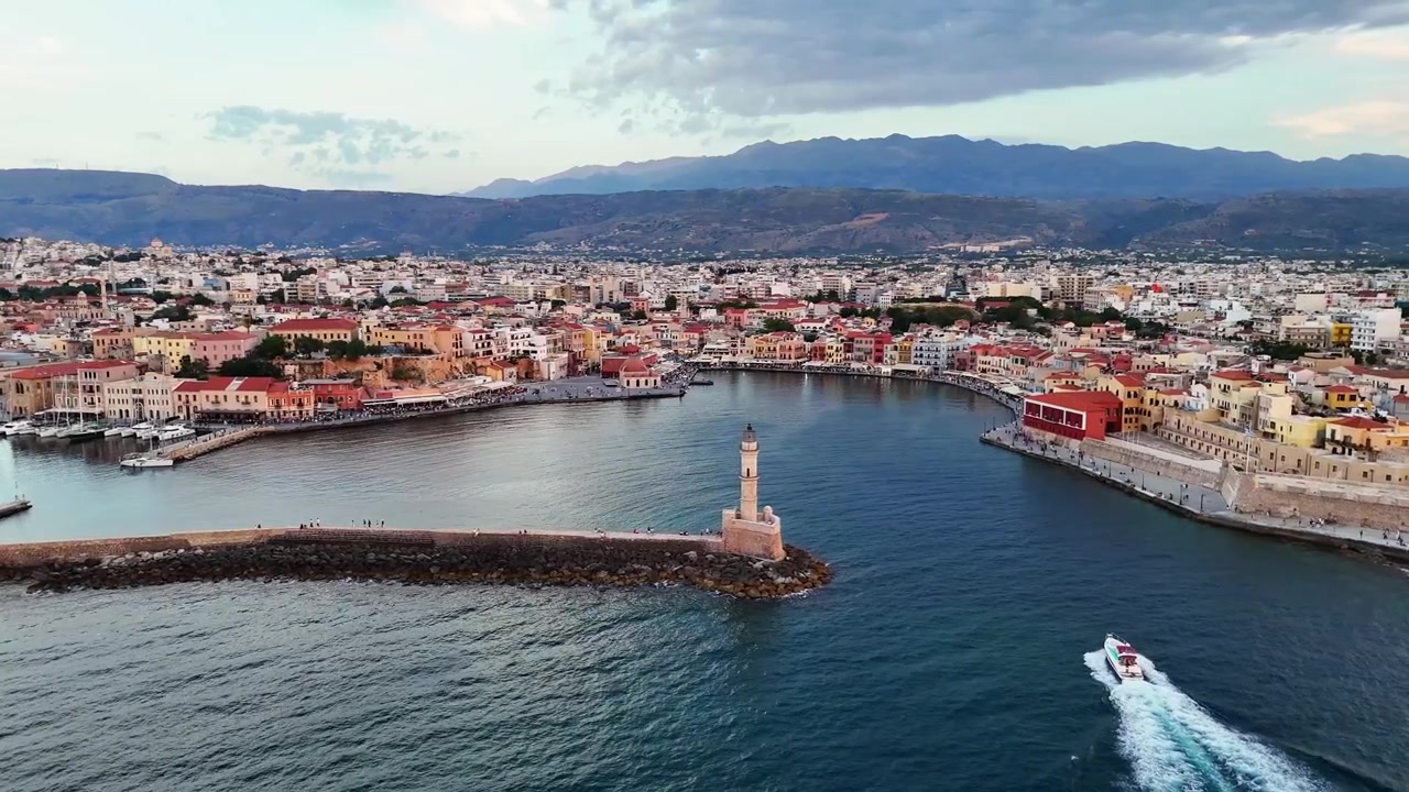 Un motoscafo entra nel porto di Chania e lascia una scia bianca accanto alla diga frangiflutti storica del faro, mentre la vista aerea mostra la città vecchia e le montagne sullo sfondo