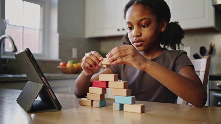 Glancing at tablet, child stacking wooden blocks into arch at kitchen table, wearing short-sleeves
