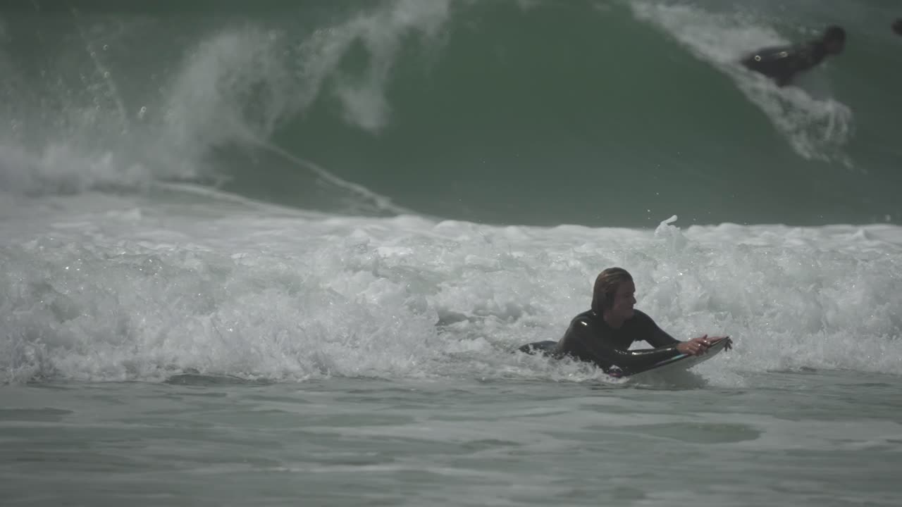 Surfer in wetsuit paddles through foamy whitewater under overcast skies, pushing toward the lineup. Realistic ocean action, endurance and focus