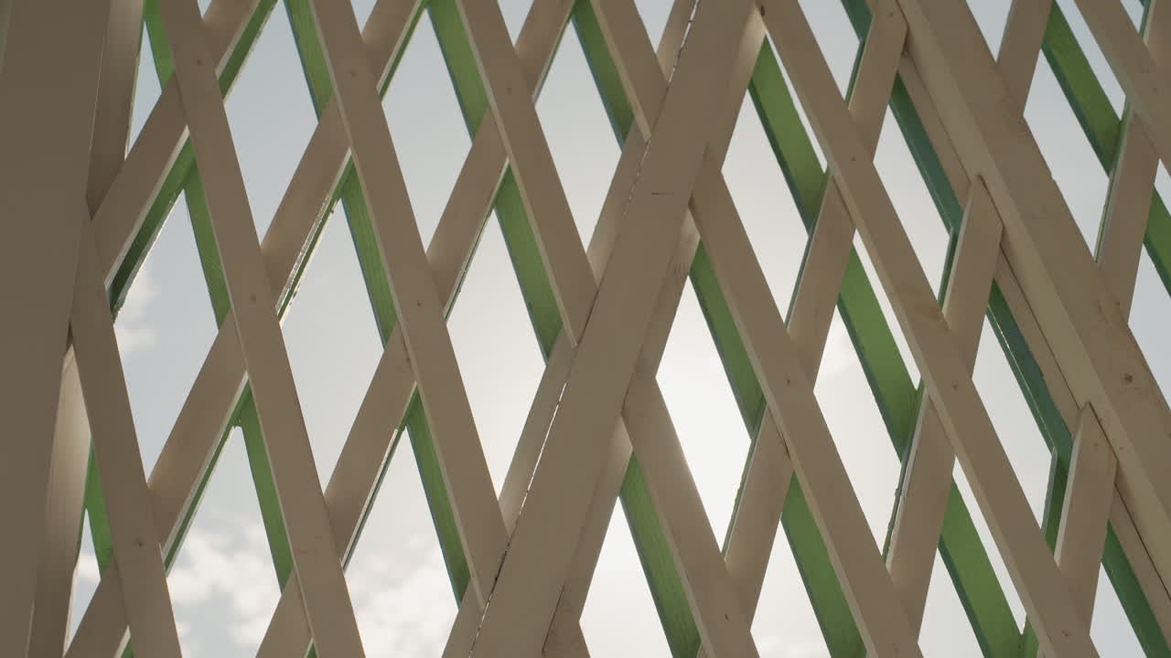 Pergola in legno illuminata dal sole come sfondo per un matrimonio, leggero bagliore solare attraverso la griglia a rombi, texture del legno calda, accenti verdi, cielo, nuvola, ombra delicata, atmosfera romantica, ambiente cerimoniale, riunione familiare.