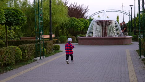 male kid wearing checkered shirt and cap runs by the green park. baby boy approaching the fountain.
