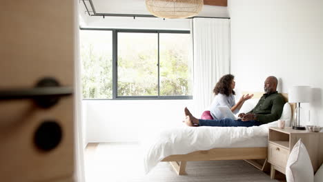 happy mature diverse couple sitting on bed, talking and using laptop, copy space, slow motion