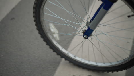 primer plano de una rueda de bicicleta moviéndose a través de un cruce de peatones marcado con rayas blancas, la rueda está en foco, mientras que una persona con pantalones grises y zapatillas de deporte camina a su lado