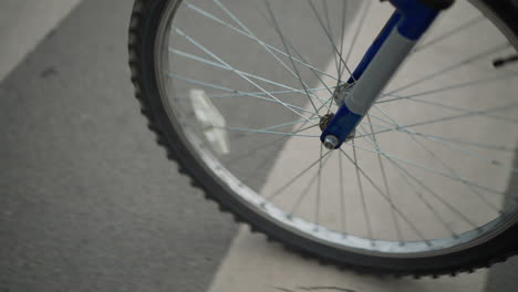 close-up of bicycle wheel moving across a pedestrian crossing marked with white stripes, the wheel is in focus, while a person wearing grey pants and sneakers walks alongside it