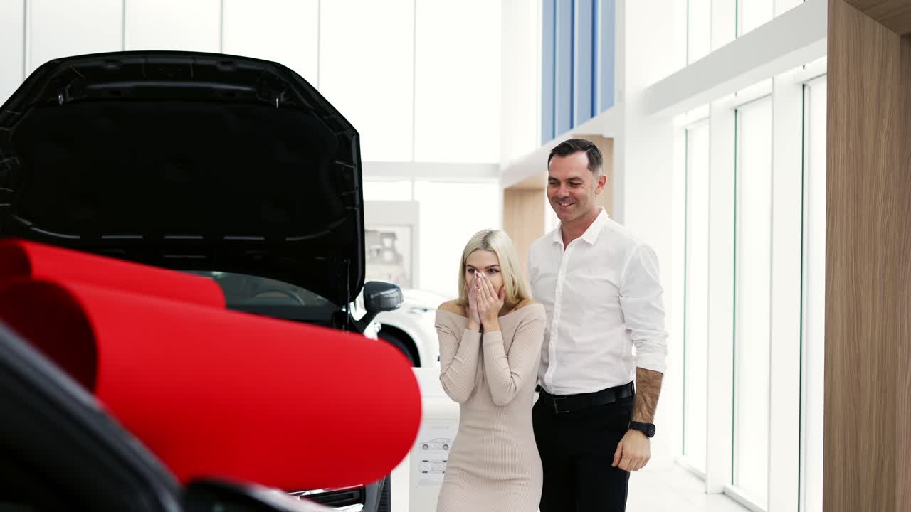 un hombre sorprende a una mujer rubia, un coche nuevo con un lazo rojo en una sala de exposiciones