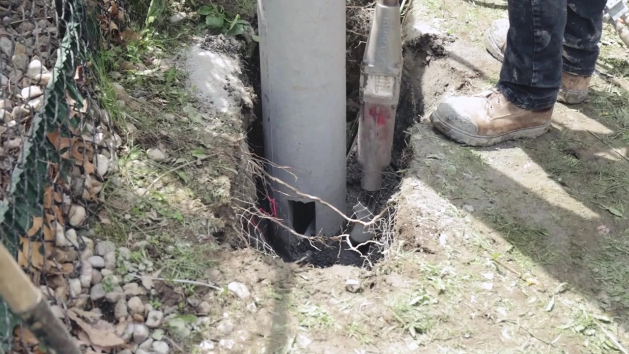 Un compattatore idraulico pressa il terreno attorno alla base di un palo di illuminazione stradale appena installato; stivali, terreno, rocce e l'attrezzo compattatore sono visibili mentre l'area della fondazione viene consolidata per stabilità
