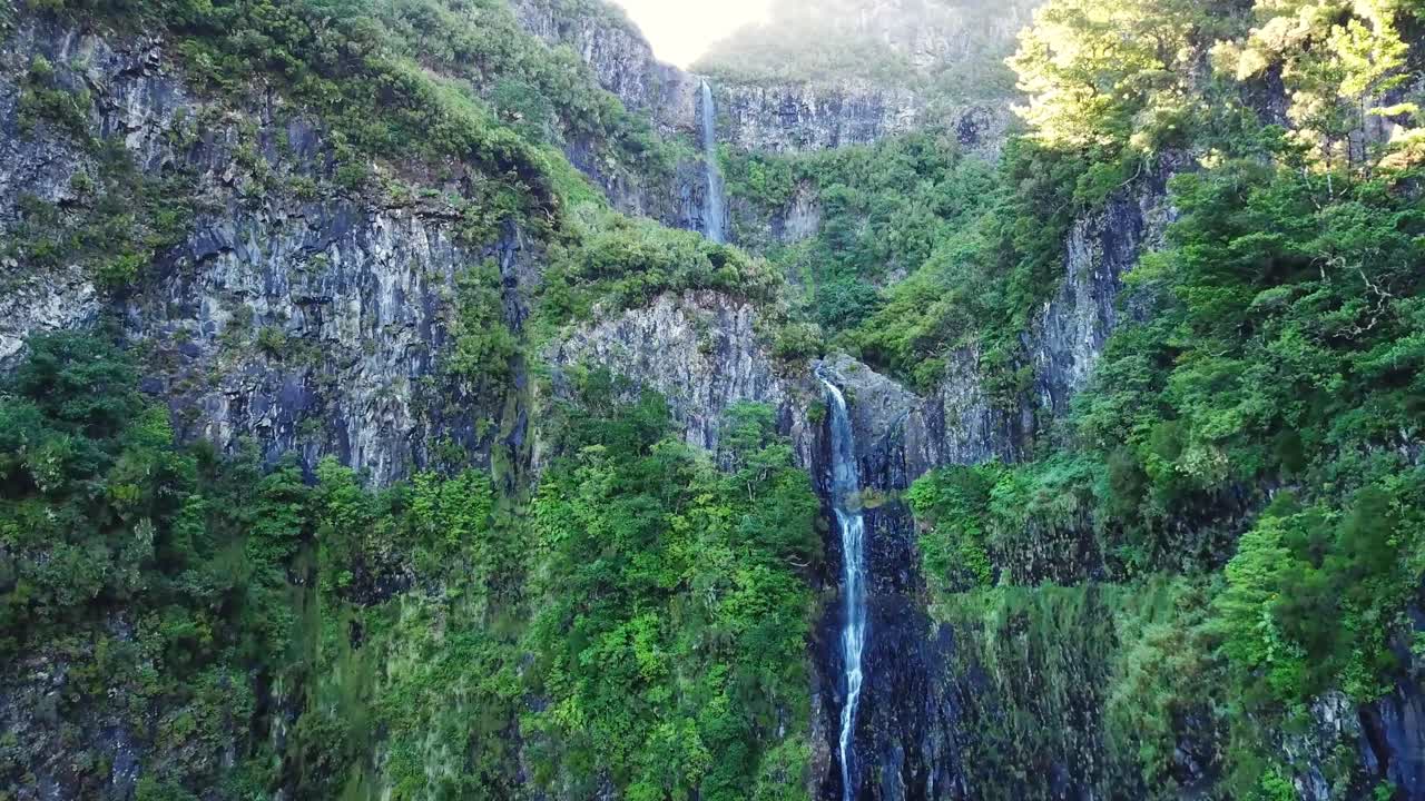 Sunlight illuminating lush green vegetation around the majestic Cascata do Risco waterfall cascading down a rocky cliff face in Madeira, Portugal, creates a stunning scene, drone flying revealing
