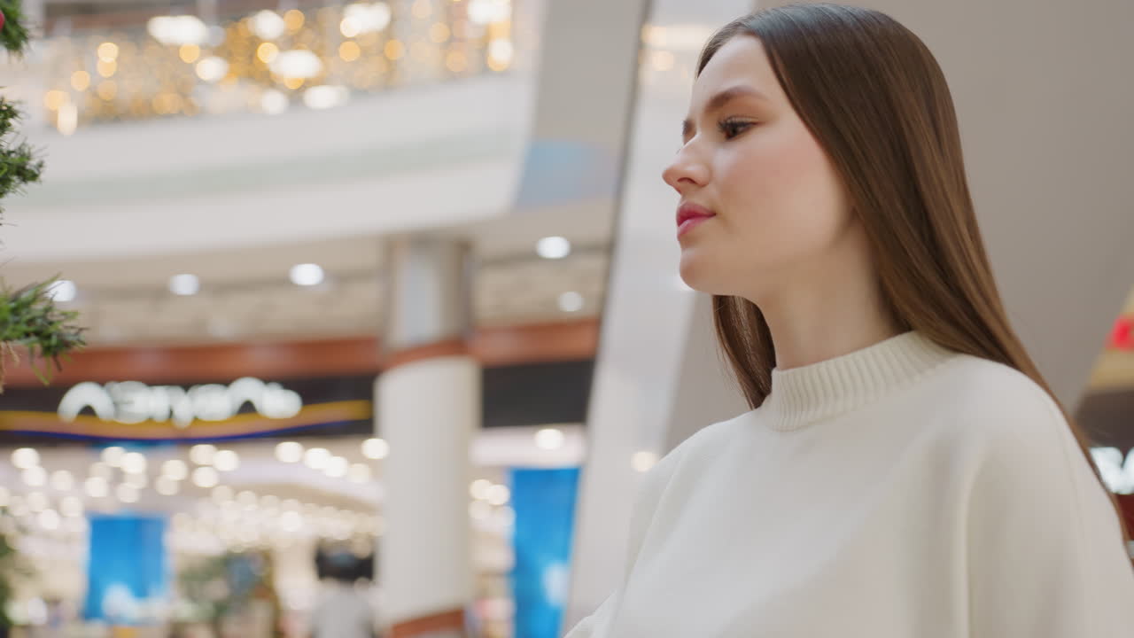 Close-up of lady smelling decorative plant in shopping mall with festive lights in background, blurred view of people enhances vibrant holiday atmosphere with warm lighting