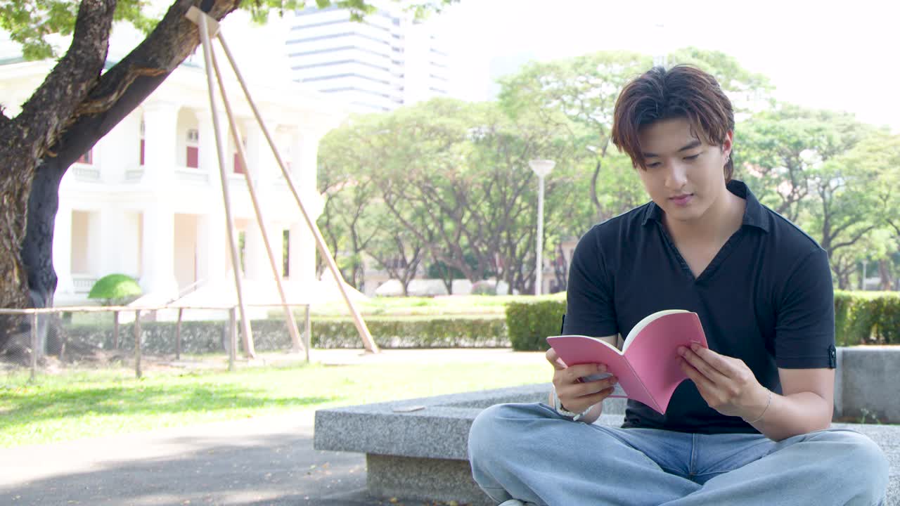 ชายหนุ่มชาวไทยนั่งขัดสมาธิอยู่บนม้านั่งหินกำลังอ่านหนังสือกลางแจ้งท่ามกลางแสงแดดจ้า