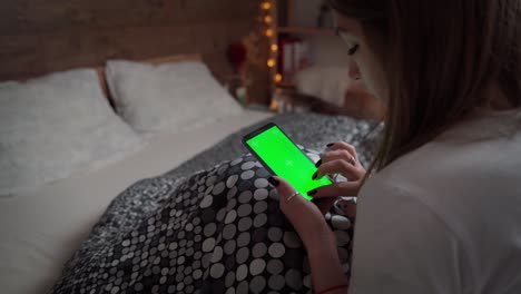mujer joven relajándose en su cama con su teléfono móvil. maqueta de niña usando la pantalla verde del teléfono inteligente. mujer hace clic con el dedo en la pantalla táctil del teléfono celular