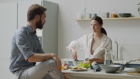 hermosa mujer mezclando ensalada en un tazón, mientras su novio sentado en un taburete le dice algo divertido y la hace reír