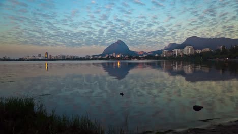 Lapso-De-Tiempo-De-Un-Amanecer-En-El-Lago-De-La-Ciudad-Rodrigo-De-Freitas-En-Río-De-Janeiro-Con-La-Orilla-En-Primer-Plano-Y-Las-Nubes-Pasando