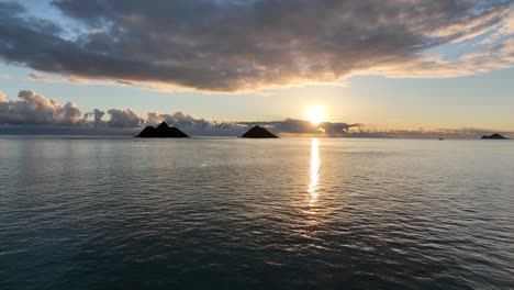 silhouette drone footage of mokulua islands at sunrise moving towards islands with clear empty water and sun rising through clouds