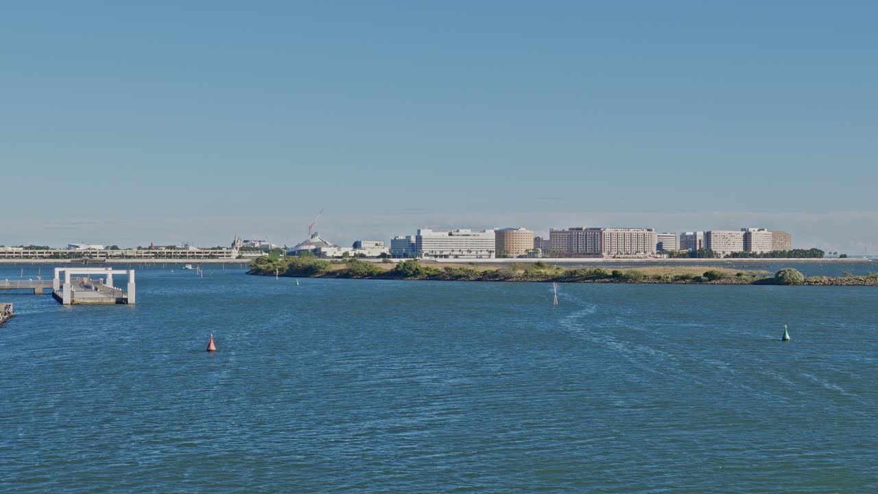 A wide view of the bay water with channel markers, showing a distant shoreline featuring large hotels or resort-style buildings and other structures