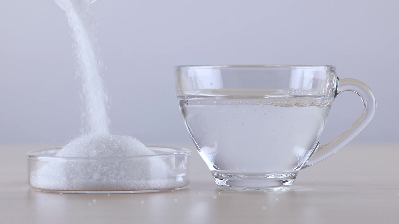 Granuli di zucchero bianco si versano in una capsula di Petri accanto a una tazza di vetro che si riempie d'acqua